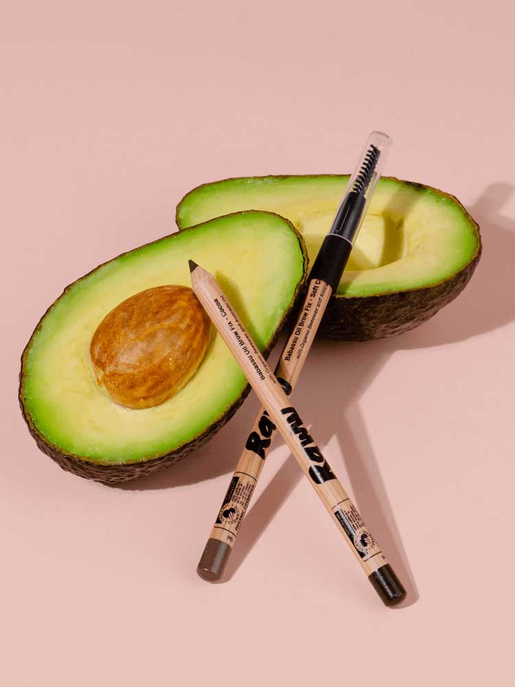 Two halves of an avocado rest on a light pink background, one with the pit. In front, two makeup pencils cross, including Raww Babassu Oil Brow Fix 1.1g for fuller-looking brows, enriched with nourishing Babassu Oil.