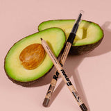 Two halves of an avocado rest on a light pink background, one with the pit. In front, two makeup pencils cross, including Raww Babassu Oil Brow Fix 1.1g for fuller-looking brows, enriched with nourishing Babassu Oil.