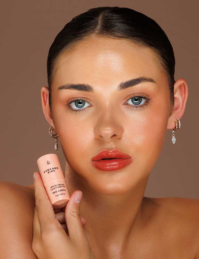 A woman with blue eyes and slicked-back hair holds Avocado Zinc SPF50 Natural Lip & Cheek Tint - Red Earth 9g near her face. Her dewy skin and glossy coral lips stand out against a warm solid background, complemented by small hoop earrings.
