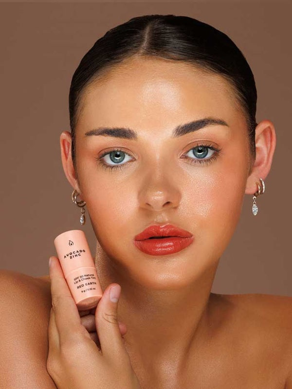 Model wearing Avocado Zinc SPF50 Natural Lip & Cheek Tint in Red Earth and holding the tint beside her face.