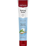 Upright white Red Seal Baking Soda Hydroxyapatite toothpaste tube with a red cap, a pale blue lower panel and mint leaves beside a wooden spoon of white powder.