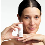 Woman holding a small white Dr Hauschka Regenerating Eye Cream bottle upright beside her cheek while resting her chin above her other hand.