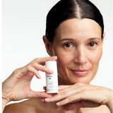 Woman holding a small white Dr Hauschka Regenerating Eye Cream bottle upright beside her cheek while resting her chin above her other hand.