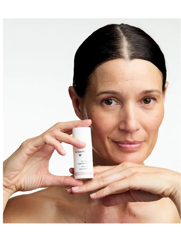 Woman holding a small white Dr Hauschka Regenerating Eye Cream bottle upright beside her cheek while resting her chin above her other hand.