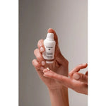 Hands holding an uncapped white Dr Hauschka Regenerating Eye Cream pump bottle, with a small white cream dab on an outstretched fingertip below it.
