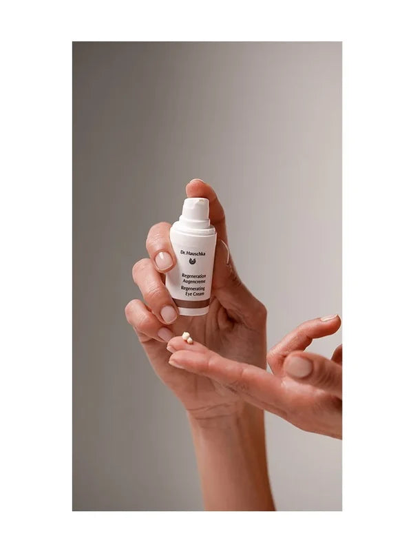 Hands holding an uncapped white Dr Hauschka Regenerating Eye Cream pump bottle, with a small white cream dab on an outstretched fingertip below it.