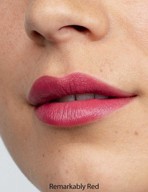 Close-up of lips wearing Antipodes Moisture Boost Natural Lipsticks 4g in matte red, showing smooth, even coverage. Lower nose and cheek are visible. Text at the bottom reads Remarkably Red.