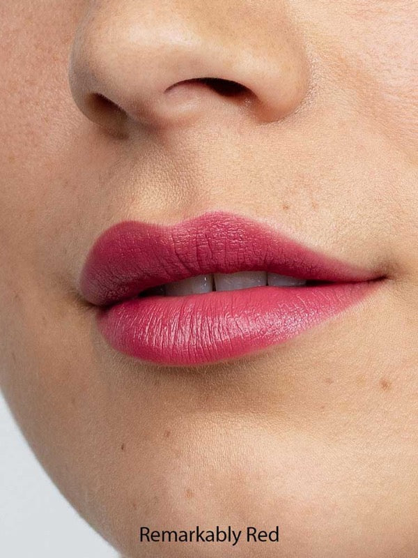 Close-up of lips wearing Antipodes Moisture Boost Natural Lipsticks 4g in matte red, showing smooth, even coverage. Lower nose and cheek are visible. Text at the bottom reads Remarkably Red.