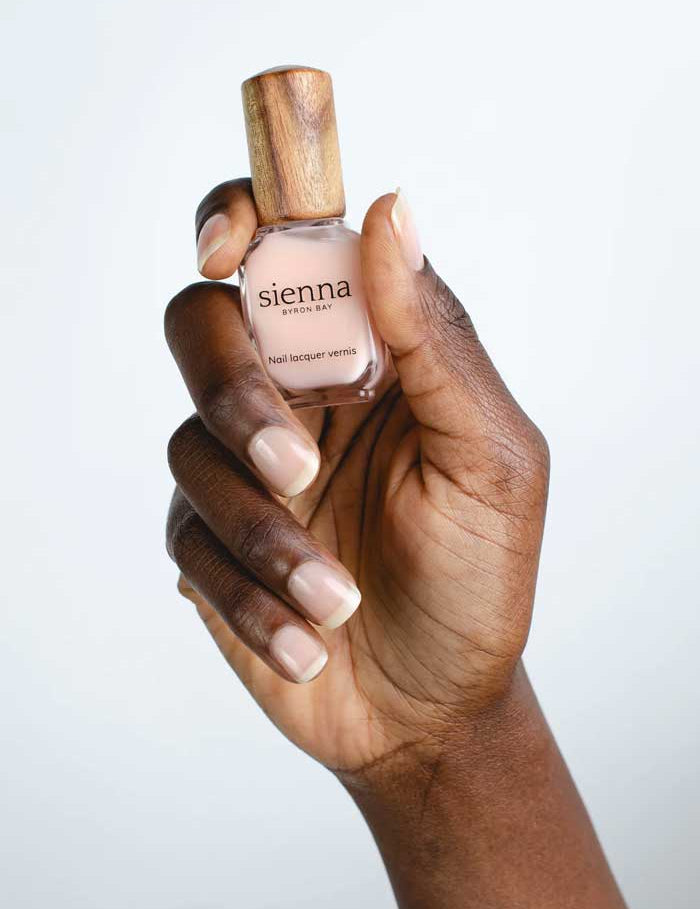 A hand with neatly manicured nails holds a bottle of Sienna Byron Bay Smooth Base Ridge Filler with a wooden cap against a plain light background.