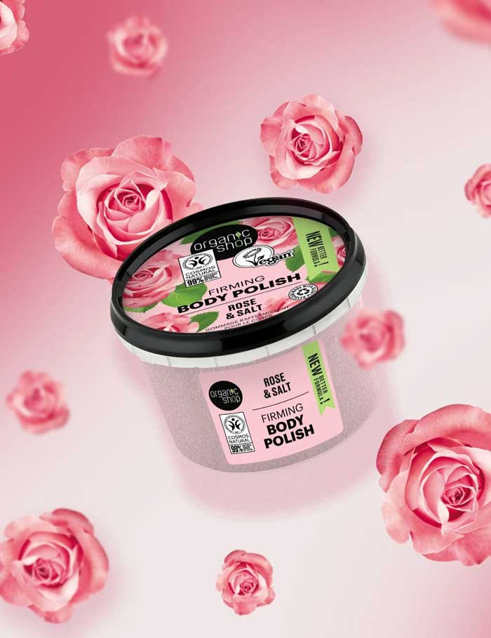 A jar of Organic Shop Rose and Salt Firming Body Polish 250ml, featuring a green, pink, and black label, is surrounded by pink roses on a pink gradient background.