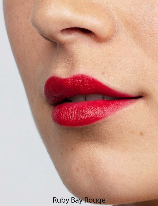 Close-up of a person's lips wearing vibrant red Antipodes Moisture Boost Natural Lipsticks 4g, with smooth skin visible around the mouth and nose. The words “Ruby Bay Rouge” appear at the bottom of the image.