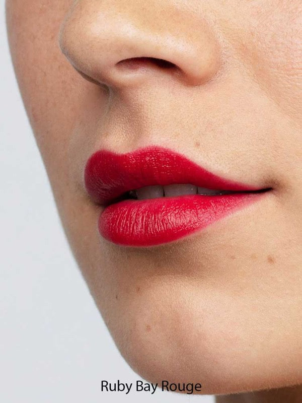 Close-up of a person's lips wearing vibrant red Antipodes Moisture Boost Natural Lipsticks 4g, with smooth skin visible around the mouth and nose. The words “Ruby Bay Rouge” appear at the bottom of the image.