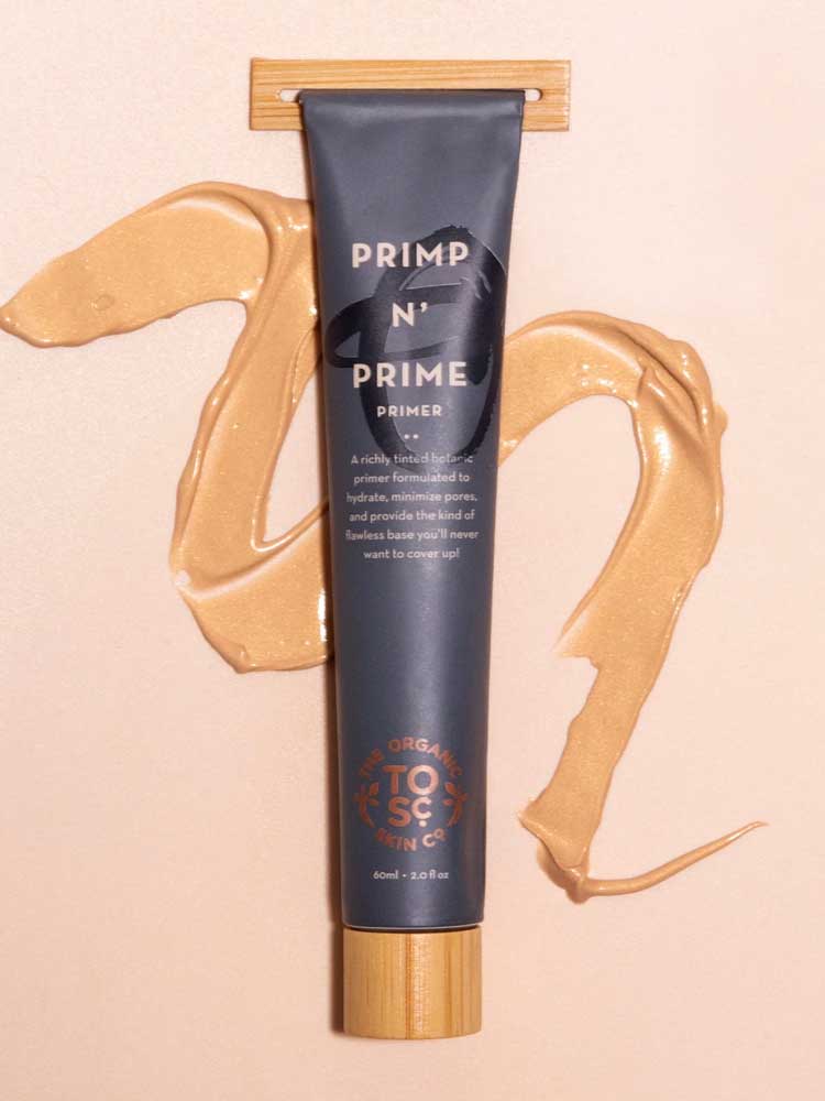 A gray tube labeled The Organic Skin Co Primp N' Prime Face Primer 60ml rests on a light surface, with beige primer cream swirled nearby. The tube has a wooden cap, gold accents, and promotes its radiant, vegan makeup primer formula.