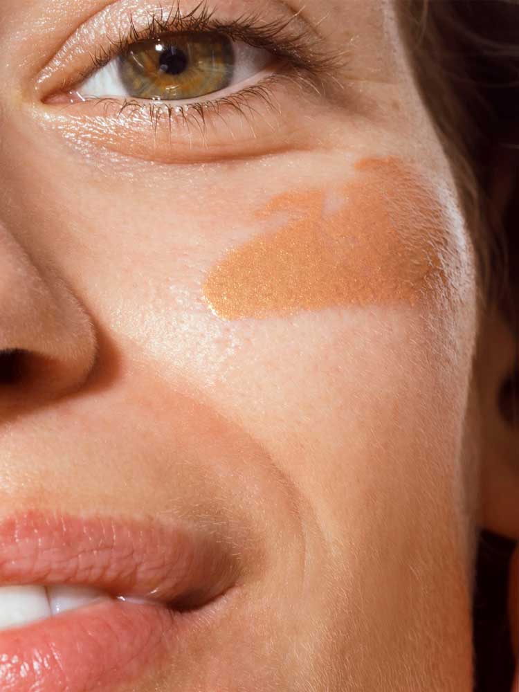 Close-up of a person’s face with brown eyes, a hint of a smile, and The Organic Skin Co Primp N' Prime Face Primer 60ml applied on their cheek. The image highlights their eye, cheek, and part of their mouth.