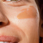 Close-up of a person’s face with brown eyes, a hint of a smile, and The Organic Skin Co Primp N' Prime Face Primer 60ml applied on their cheek. The image highlights their eye, cheek, and part of their mouth.