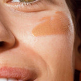 Close-up of a person’s face with brown eyes, a hint of a smile, and The Organic Skin Co Primp N' Prime Face Primer 60ml applied on their cheek. The image highlights their eye, cheek, and part of their mouth.