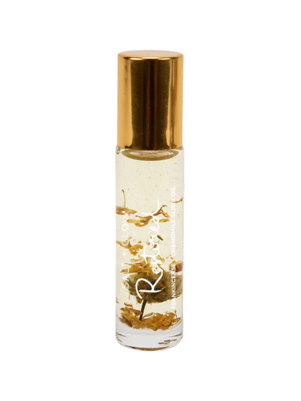 Slim clear Salt + Glow Restored roll-on perfume bottle with a shiny gold cap, pale golden liquid and dried golden-brown botanical pieces inside.