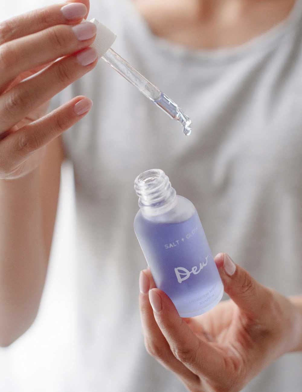 A person wearing a light gray shirt holds a dropper above an open, frosted glass bottle labeled Salt + Glow Dew Hydration Serum 30ml, filled with purple serum for dry skin, ready to dispense a drop.