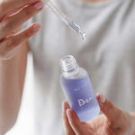 A person wearing a light gray shirt holds a dropper above an open, frosted glass bottle labeled Salt + Glow Dew Hydration Serum 30ml, filled with purple serum for dry skin, ready to dispense a drop.