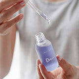 A person wearing a light gray shirt holds a dropper above an open, frosted glass bottle labeled Salt + Glow Dew Hydration Serum 30ml, filled with purple serum for dry skin, ready to dispense a drop.