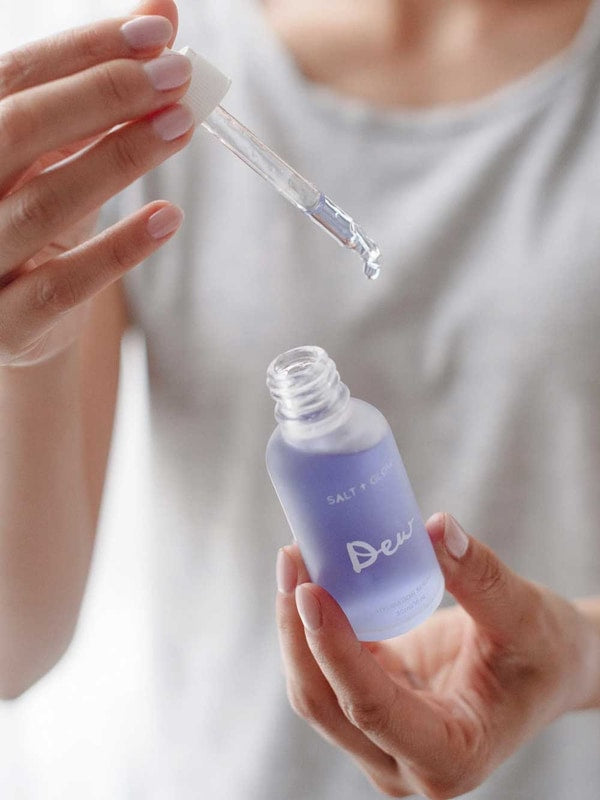 A person wearing a light gray shirt holds a dropper above an open, frosted glass bottle labeled Salt + Glow Dew Hydration Serum 30ml, filled with purple serum for dry skin, ready to dispense a drop.