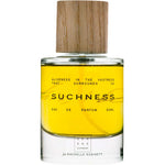 A clear glass bottle of Serene Body Health Natural Eau de Parfum Suchness 50ml, vegan and yellow, with a wooden cap. The label reads: Aliveness in the vastness that surrounds us, natural eau de parfum 50ml, for Rachelle Robinett.