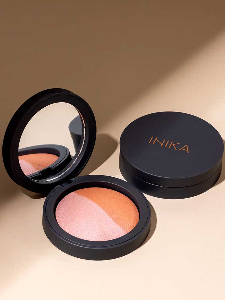 An open INIKA Organic Mineral Baked Blush Duo - Burnt Peach 6.5g with a mirror displays pink and bronze shades beside its closed compact, both set on a beige surface.