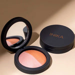 An open INIKA Organic Mineral Baked Blush Duo - Burnt Peach 6.5g with a mirror displays pink and bronze shades beside its closed compact, both set on a beige surface.
