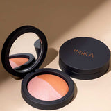 An open INIKA Organic Mineral Baked Blush Duo - Burnt Peach 6.5g with a mirror displays pink and bronze shades beside its closed compact, both set on a beige surface.