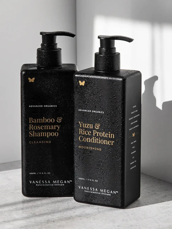 Black rectangular Vanessa Megan Yuzu & Rice Protein Conditioner and Bamboo & Rosemary Shampoo pump bottles stand together in sunlight, with water droplets on their surfaces.