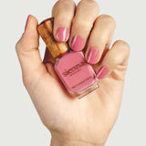 A hand with neatly manicured pink nails holds a bottle of Sienna Byron Bay - Blossom nail polish, featuring a wooden cap against a light gray background.