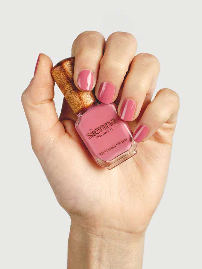 A hand with neatly manicured pink nails holds a bottle of Sienna Byron Bay - Blossom nail polish, featuring a wooden cap against a light gray background.