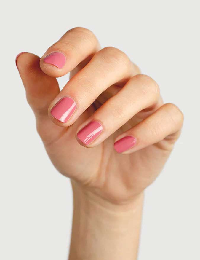 A close-up of a hand with neatly manicured nails painted in Sienna Byron Bay's Blossom, a glossy pink polish, set against a plain light gray background.