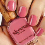 A hand with neatly manicured nails painted pink holds a bottle of Sienna Byron Bay - Blossom nail polish, featuring a wooden cap and elegant minimalist branding.