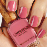 A hand with neatly manicured nails painted pink holds a bottle of Sienna Byron Bay - Blossom nail polish, featuring a wooden cap and elegant minimalist branding.