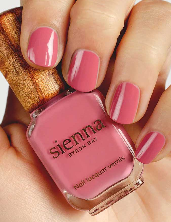 A hand with neatly manicured nails painted pink holds a bottle of Sienna Byron Bay - Blossom nail polish, featuring a wooden cap and elegant minimalist branding.