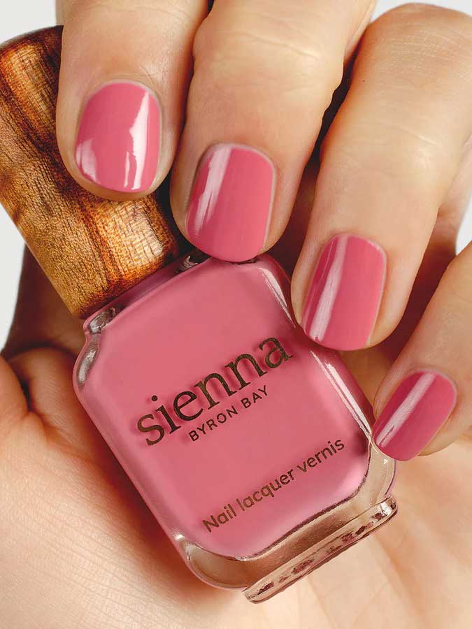 A hand with neatly manicured nails painted pink holds a bottle of Sienna Byron Bay - Blossom nail polish, featuring a wooden cap and elegant minimalist branding.