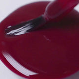 A close-up shows Sienna Byron Bay Nail Polish - Promise, a deep burgundy red, being brushed onto a white surface, forming a glossy puddle.