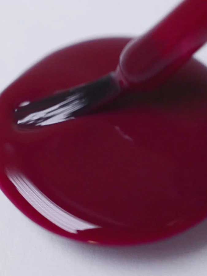 A close-up shows Sienna Byron Bay Nail Polish - Promise, a deep burgundy red, being brushed onto a white surface, forming a glossy puddle.