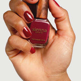 A hand with neatly manicured nails painted deep red holds a bottle of Sienna Byron Bay Nail Polish - Promise against a plain background.