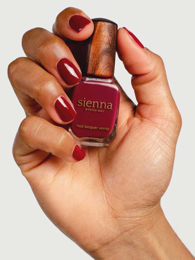 A hand with neatly manicured nails painted deep red holds a bottle of Sienna Byron Bay Nail Polish - Promise against a plain background.