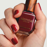 A hand with neatly manicured nails painted deep red holds a bottle of Sienna Byron Bay Nail Polish in the shade "Promise," featuring the brand's signature wooden cap.