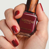 A hand with neatly manicured nails painted deep red holds a bottle of Sienna Byron Bay Nail Polish in the shade "Promise," featuring the brand's signature wooden cap.