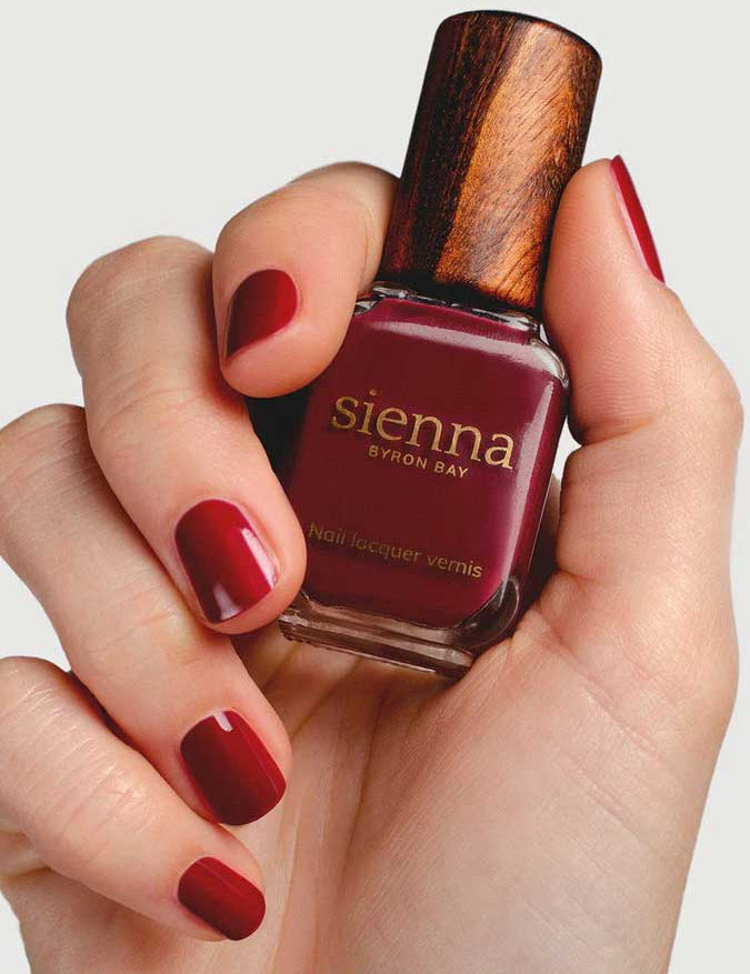 A hand with neatly manicured nails painted deep red holds a bottle of Sienna Byron Bay Nail Polish in the shade "Promise," featuring the brand's signature wooden cap.