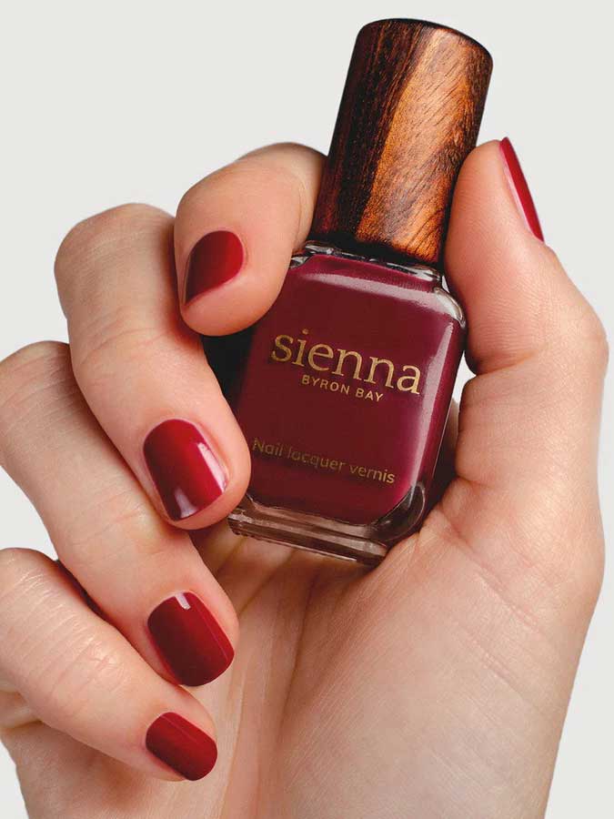 A hand with neatly manicured nails painted deep red holds a bottle of Sienna Byron Bay Nail Polish in the shade "Promise," featuring the brand's signature wooden cap.