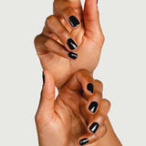 Two hands with dark skin, fingers gently intertwined, display short nails polished in glossy black using Sienna Byron Bay Nail Polish in Onyx by Sienna Byron Bay against a plain background.