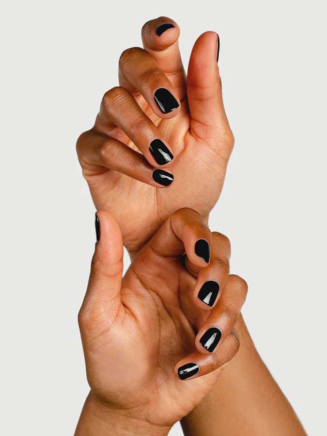 Two hands with dark skin, fingers gently intertwined, display short nails polished in glossy black using Sienna Byron Bay Nail Polish in Onyx by Sienna Byron Bay against a plain background.