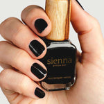 A hand with neatly manicured black nails holds a bottle of Sienna Byron Bay Nail Polish in the shade Onyx, featuring a wooden cap, against a plain light background.