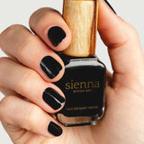 A hand with neatly manicured black nails holds a bottle of Sienna Byron Bay Nail Polish in the shade Onyx, featuring a wooden cap, against a plain light background.