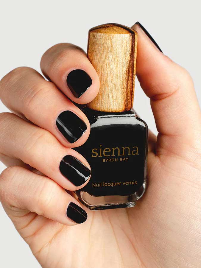 A hand with neatly manicured black nails holds a bottle of Sienna Byron Bay Nail Polish in the shade Onyx, featuring a wooden cap, against a plain light background.